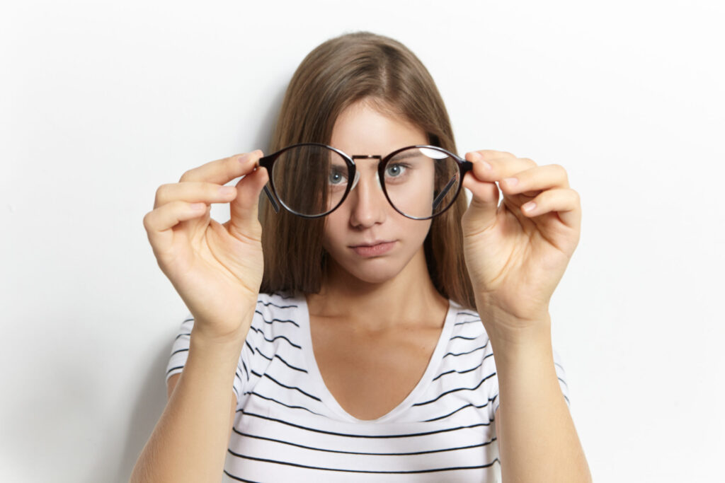 Beautiful cute shortsighted teeage girl in stylish striped t-shirt putting on her first eyeglasses. Vision correction, optics, nearsightedness and myopia concept. Selective focus on spectacles
